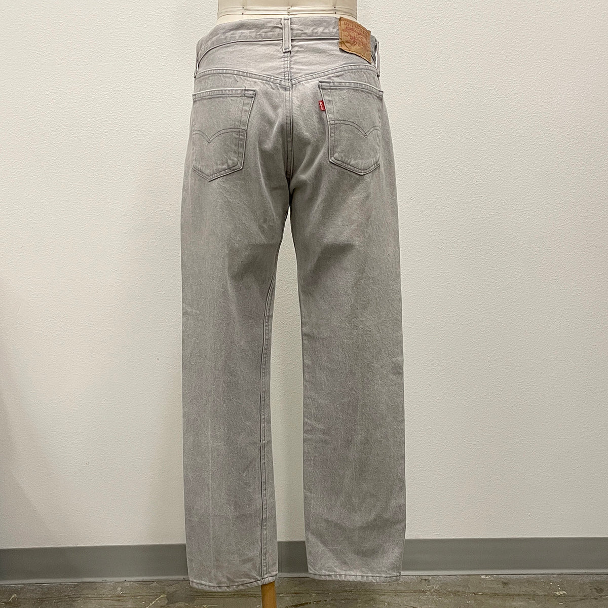 Light gray washed denim jeans shown from the back on a mannequin, featuring a brown leather patch on the back waistband and a small red tab on the back pocket.
