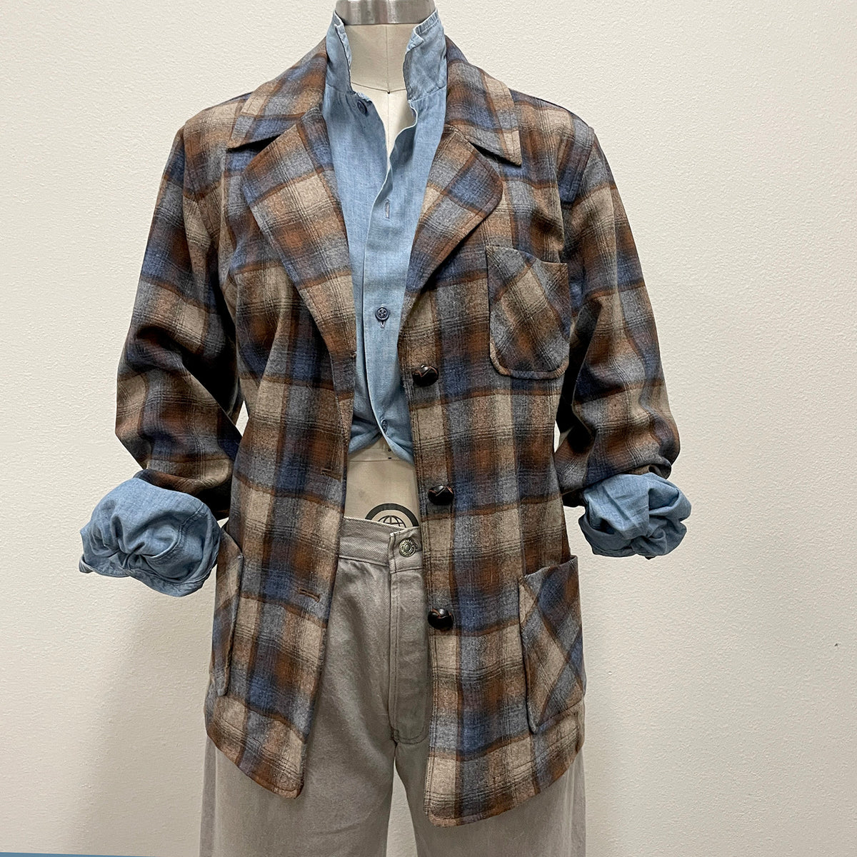 A vintage plaid blazer jacket in brown, beige, and blue tones, styled on a mannequin with a light blue button-down shirt and grey jeans against a plain off-white wall.