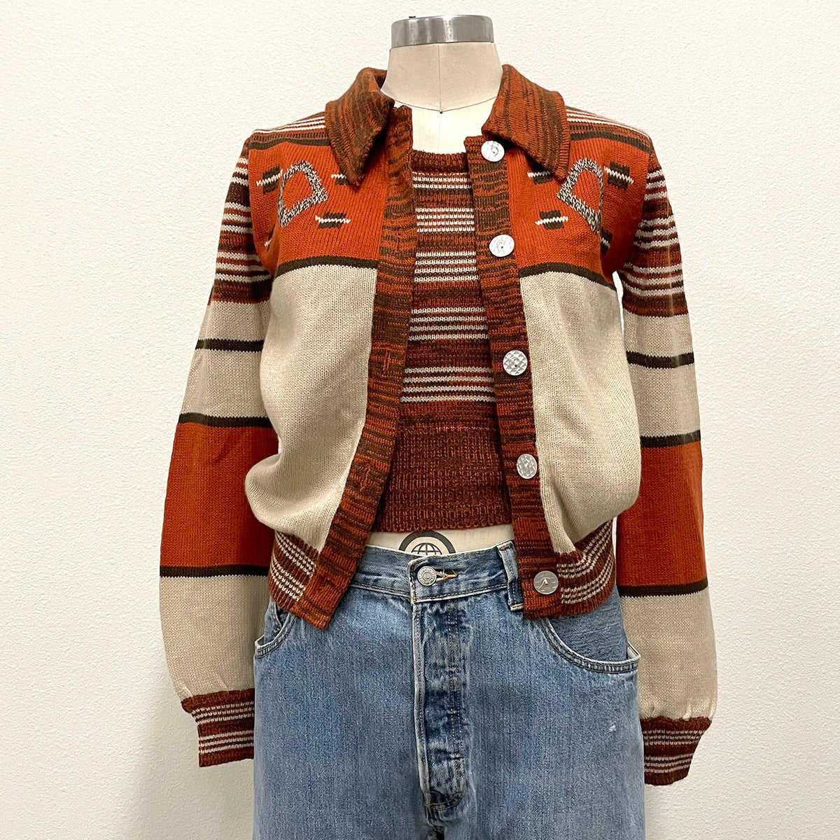 A vintage knit cardigan sweater displayed on a mannequin torso, paired with a striped knit top and blue jeans. The cardigan features a warm rust orange, cream, and brown color-blocked design with geometric pattern on the chest and pearl-tone buttons.