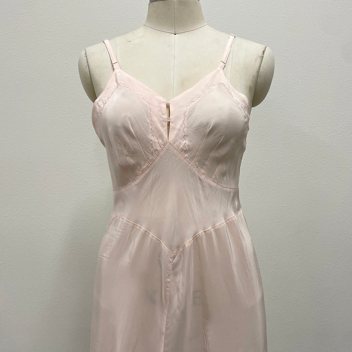 A pale blush pink silky slip dress with thin spaghetti straps and a V-neckline, displayed on a cream-colored mannequin torso against a plain light gray wall.