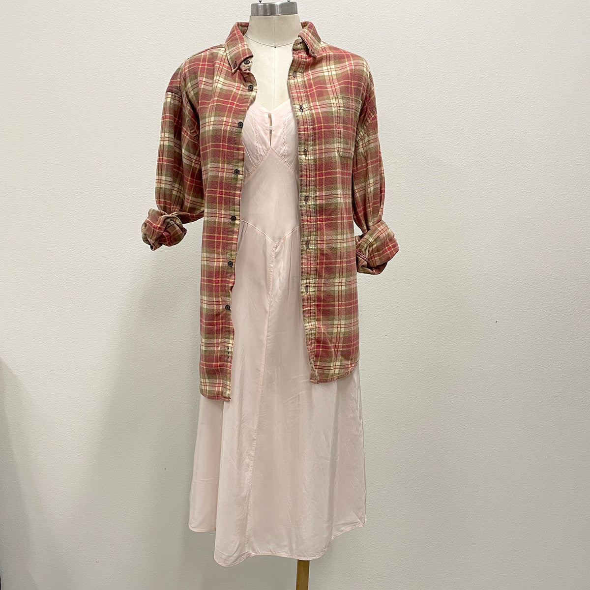 A long-sleeve red and beige plaid flannel shirt styled over a pink silk slip dress on a mannequin against a plain white wall.