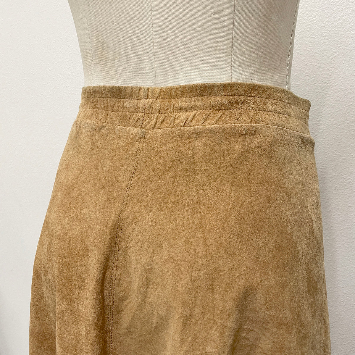 A close-up of a tan suede skirt displayed on a cream dress form against a white wall.
