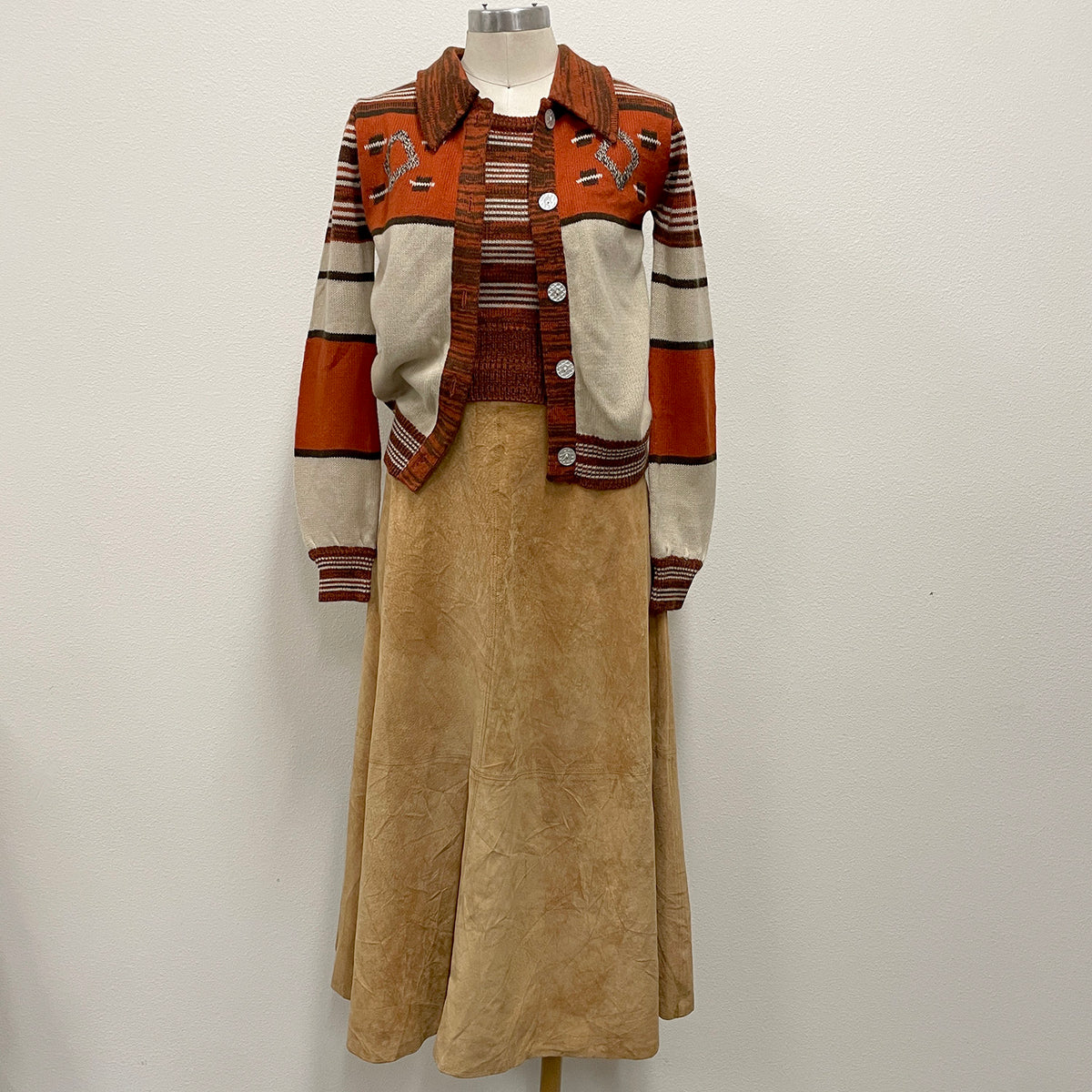 A vintage-style two-piece knit set displayed on a mannequin, featuring a long-sleeve button-front cardigan with western-inspired stripes and embroidery, paired with a tan suede a-line, maxi skirt.