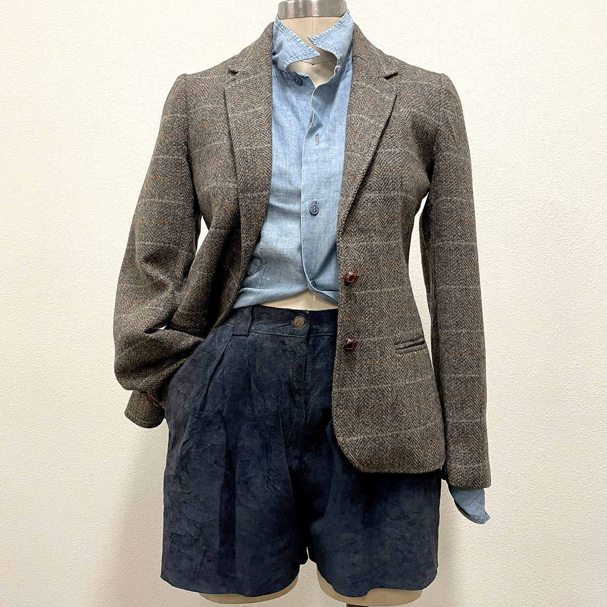 A two-piece suit set displayed on a mannequin, featuring a brown and gray windowpane plaid blazer over a light blue button-down shirt, paired with navy suede shorts.