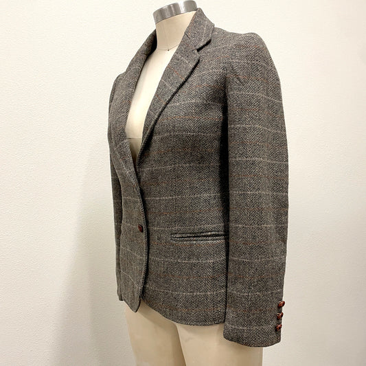 A gray and brown herringbone tweed blazer with a subtle windowpane check pattern, displayed on a cream-colored mannequin torso against a plain off-white wall.