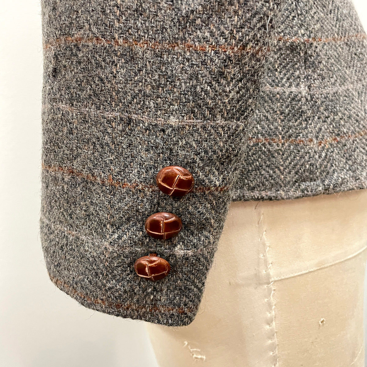 Close-up of a gray and black herringbone tweed blazer sleeve with three brown leather buttons, displayed on a cream-colored mannequin torso.