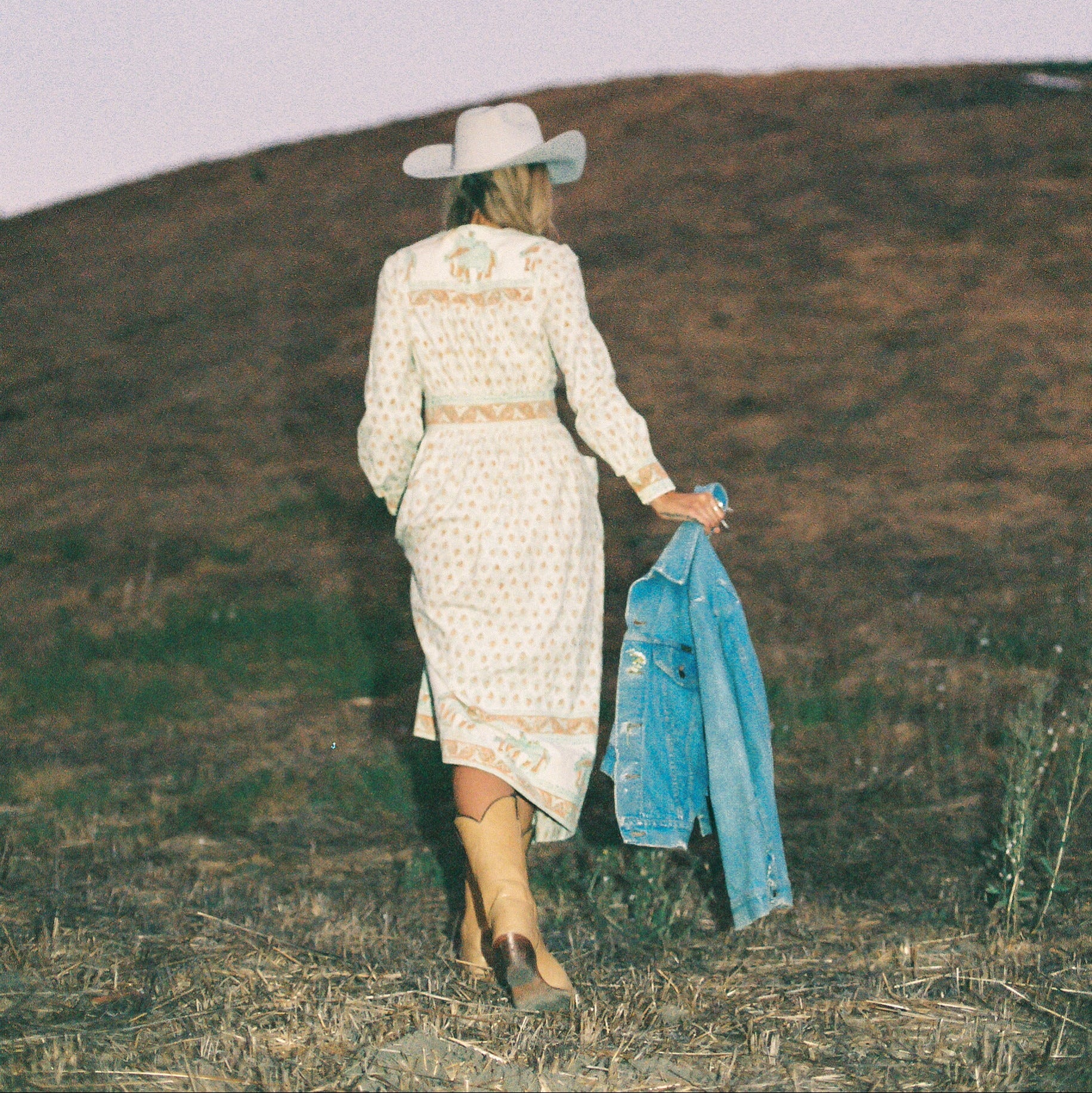 Woman in a floral print dress and cowboy hat holding a denim jacket on a grassy hill.