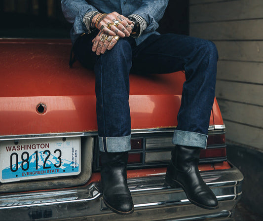 Man in a hat sitting on the back of a red car in Dark selvage jeans with a dark background