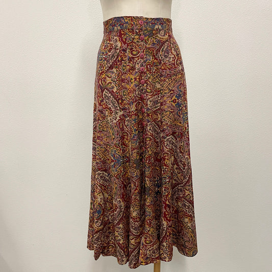 A high-waisted, full skirt with an intricate paisley pattern in red, brown, gold, and blue, displayed on a mannequin torso against a plain white wall.
