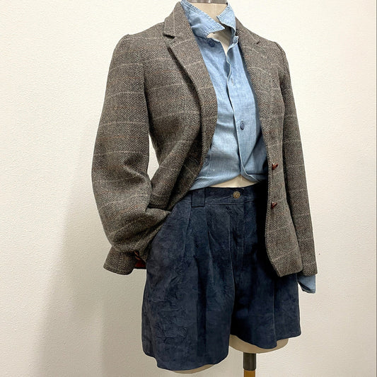 A gray and brown plaid tweed blazer styled on a mannequin with a light blue button-down shirt and dark blue pleated shorts against a plain off-white wall.