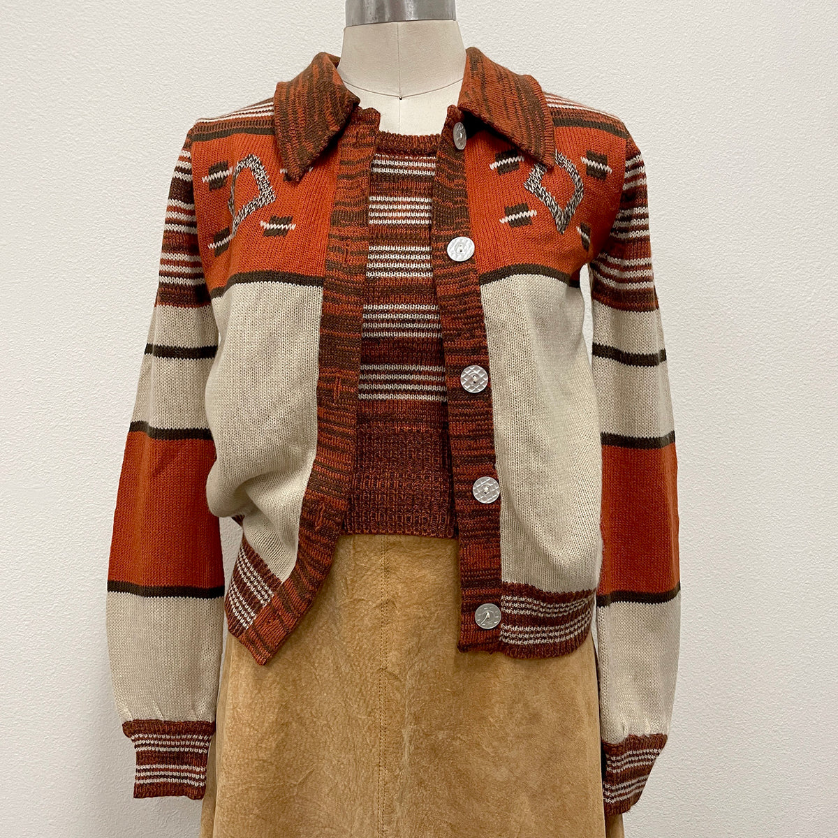 A vintage knit cardigan sweater displayed on a mannequin torso, featuring a rust orange, cream, and brown striped design with decorative intarsia details on the chest and a button-up front.