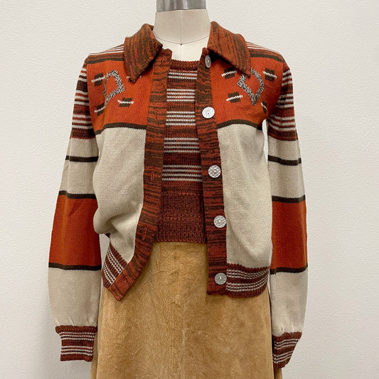 A vintage knit cardigan sweater displayed on a mannequin torso, featuring a rust orange, cream, and brown striped design with decorative intarsia details on the chest and a button-up front.