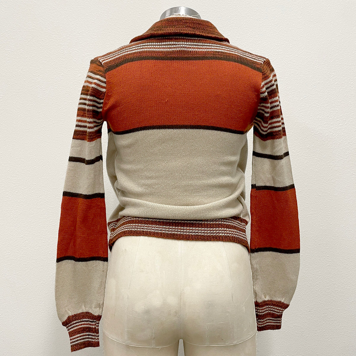 A striped knit sweater displayed on a cream-colored mannequin torso against a plain off-white background. The sweater features a rust orange body with horizontal stripes in brown, rust, and cream on the sleeves and collar.