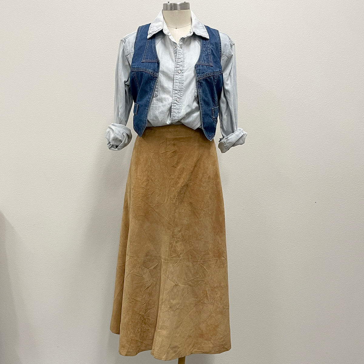 A three-piece outfit displayed on a mannequin, featuring a light blue denim shirt with rolled sleeves, a blue denim vest, and a tan suede skirt.