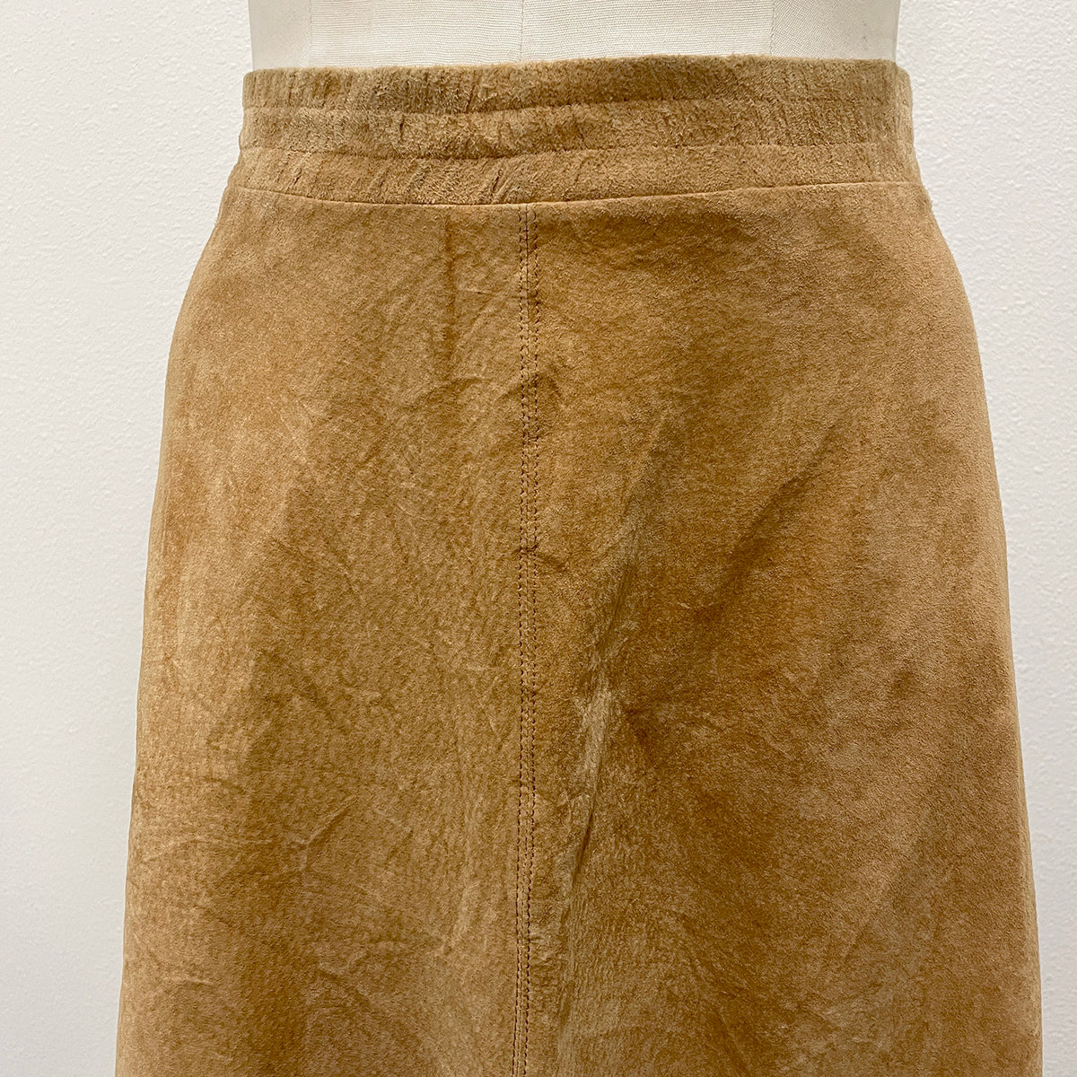 A close-up of a tan suede skirt displayed on a mannequin torso against a plain off-white wall.