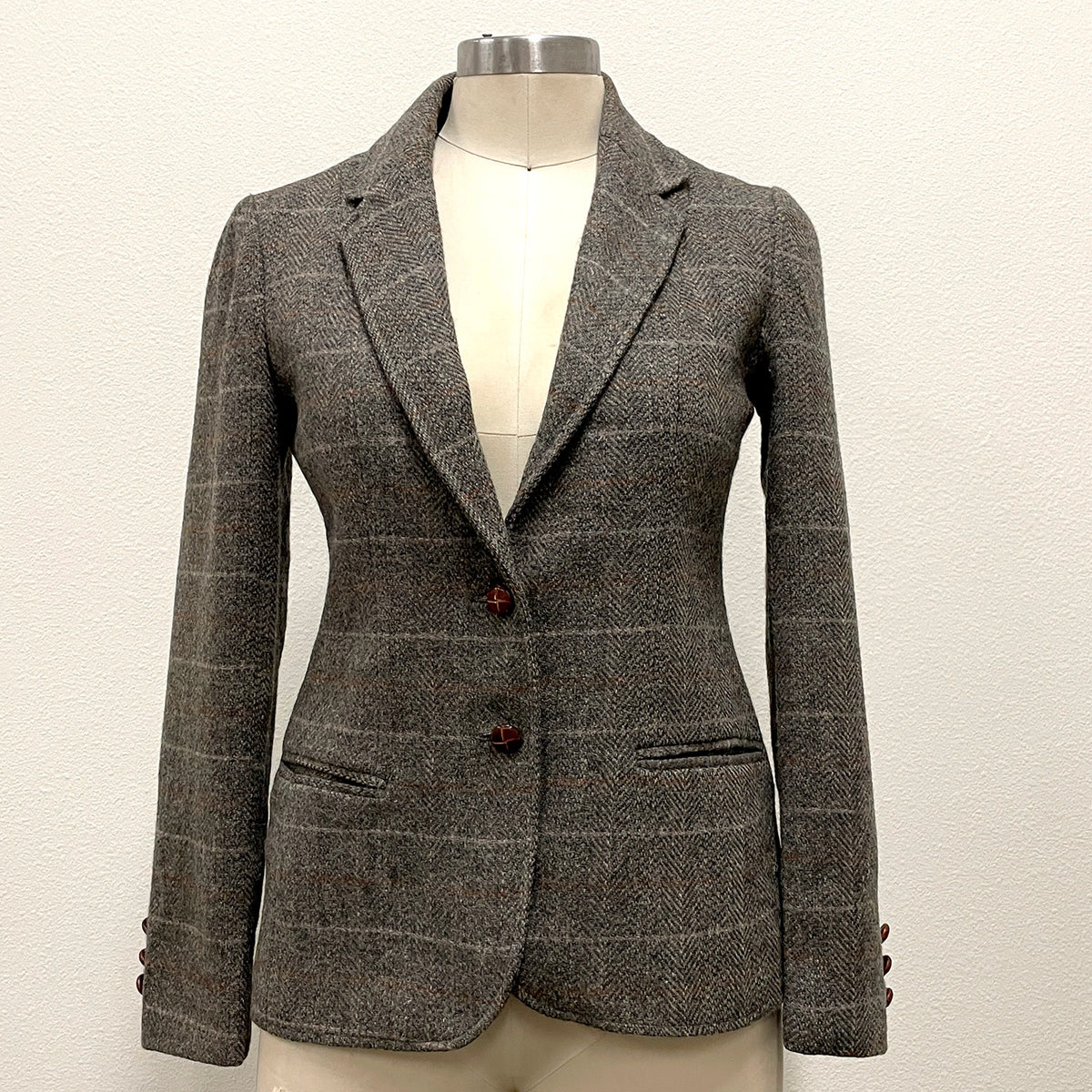 A gray and brown tweed blazer with a subtle windowpane check pattern, displayed on a cream-colored mannequin torso. The blazer features notch lapels, a two-button front closure, and welt pockets.