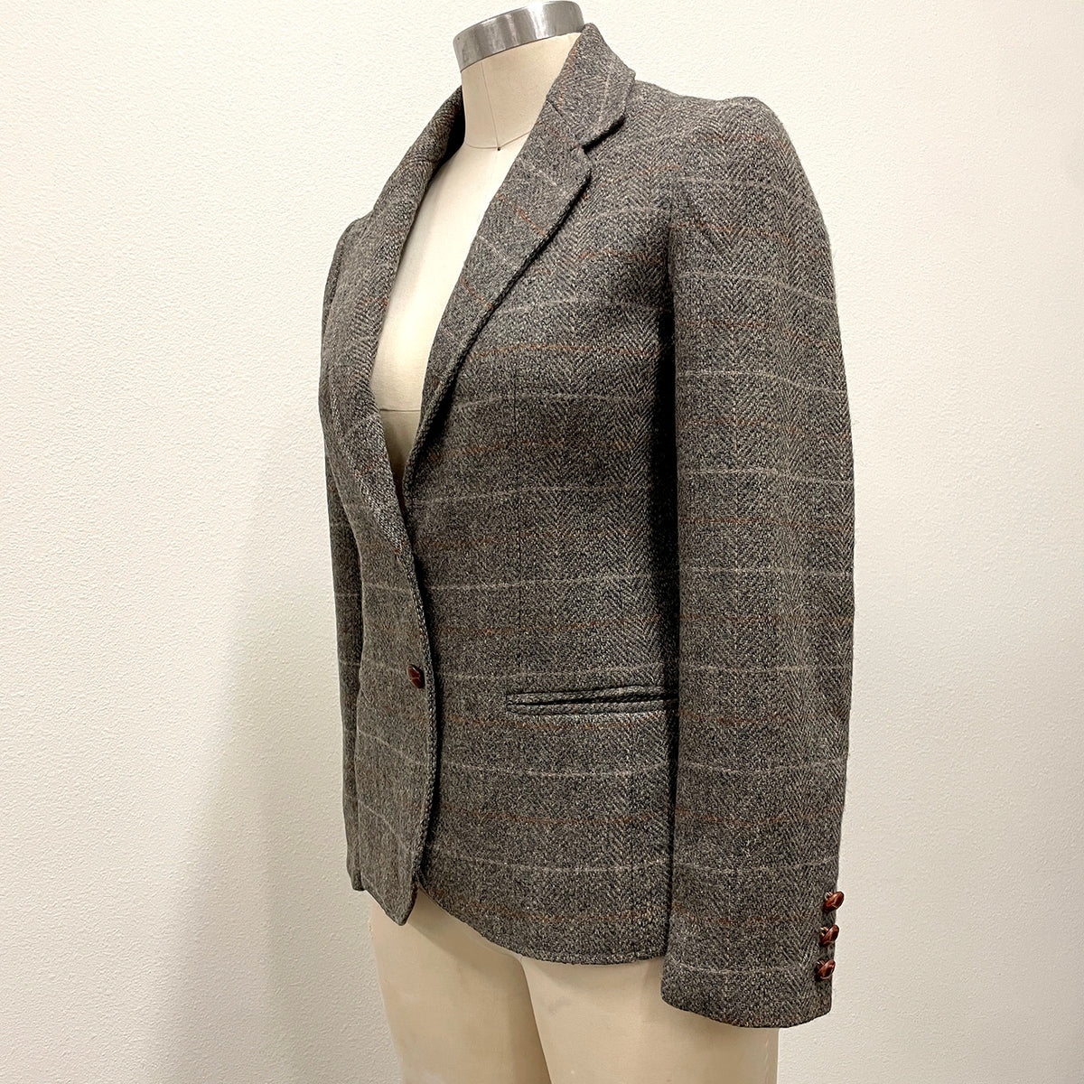 A gray and brown herringbone tweed blazer with a subtle windowpane check pattern, displayed on a cream-colored mannequin torso against a plain off-white wall.