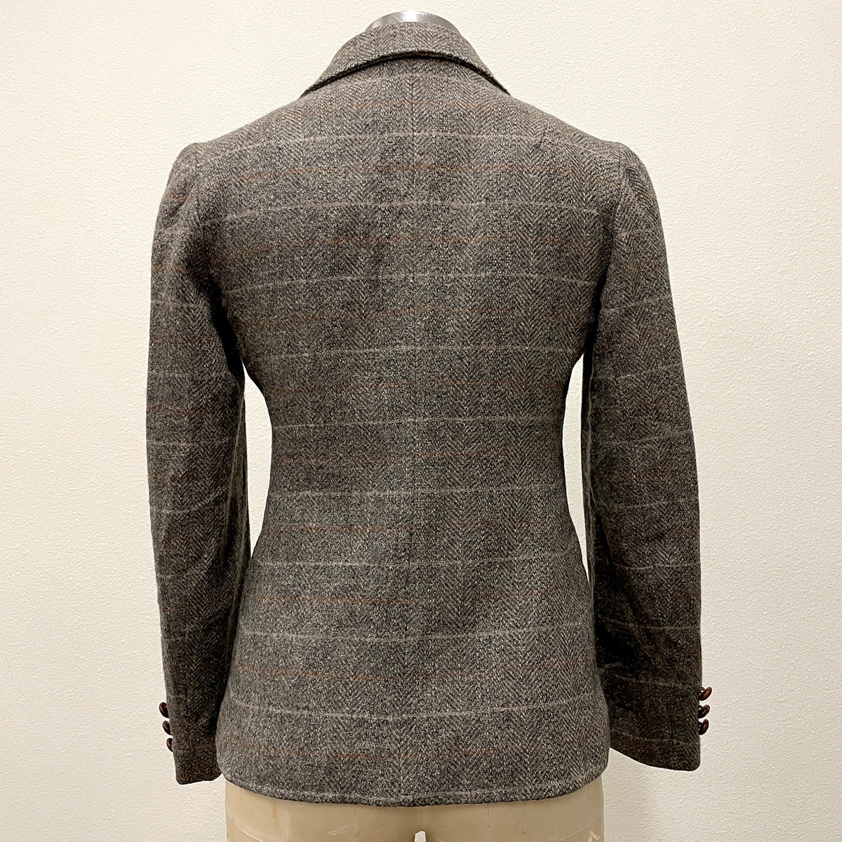 A gray and brown herringbone tweed blazer with a subtle windowpane check pattern, shown from the back on a mannequin against a plain off-white wall.